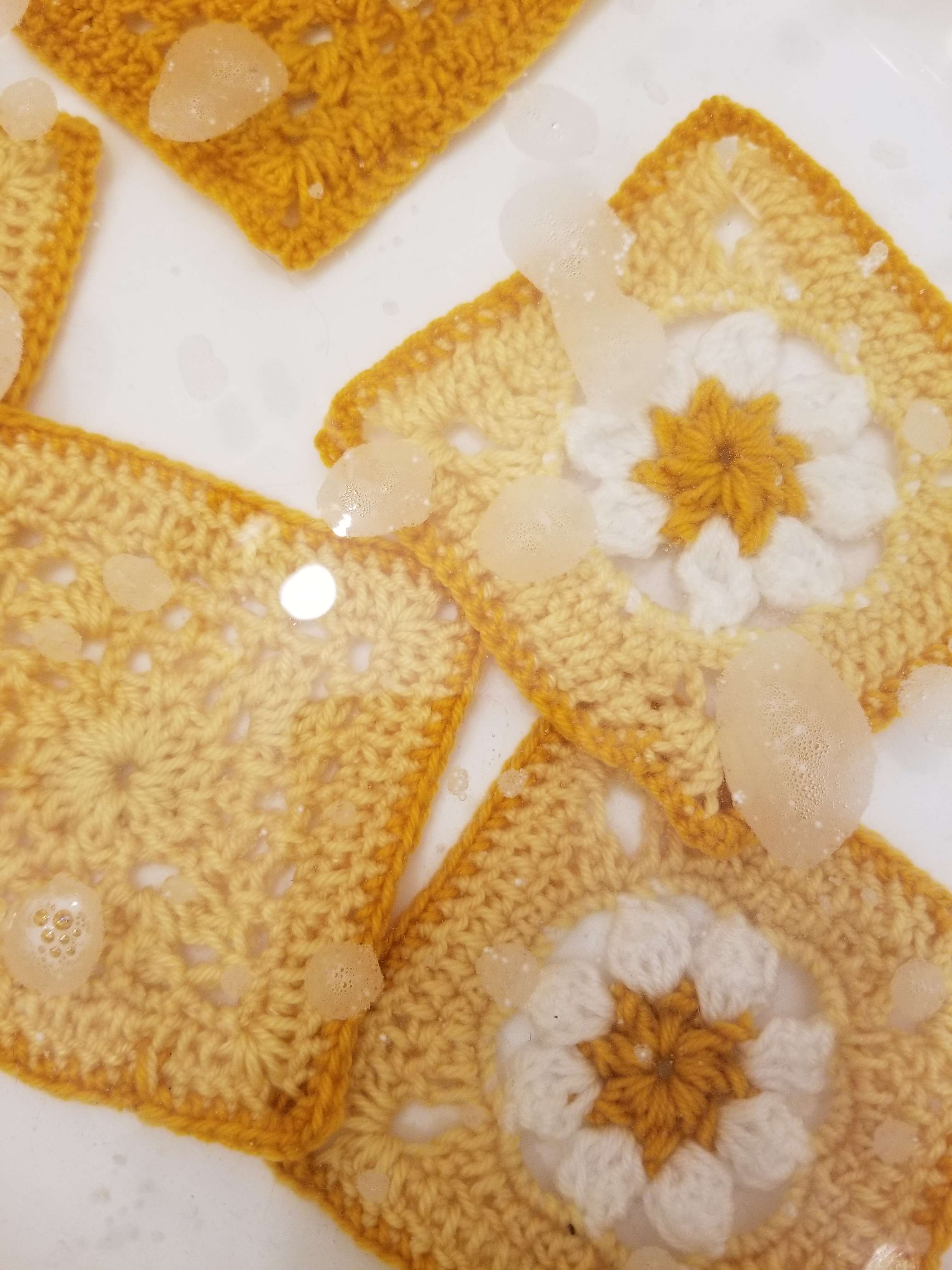 A photograph of yellow crocheted squares with a daisy motif soaking in a tub of soapy water.