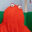 A bright red humanoid uppet with soulless eyes resting on top of his mop-like head of string.