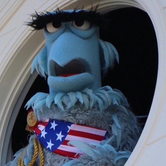 A blue eagle puppet with very large, dark eyebrows and a fluffy ruff of light blue feathers around his neck.